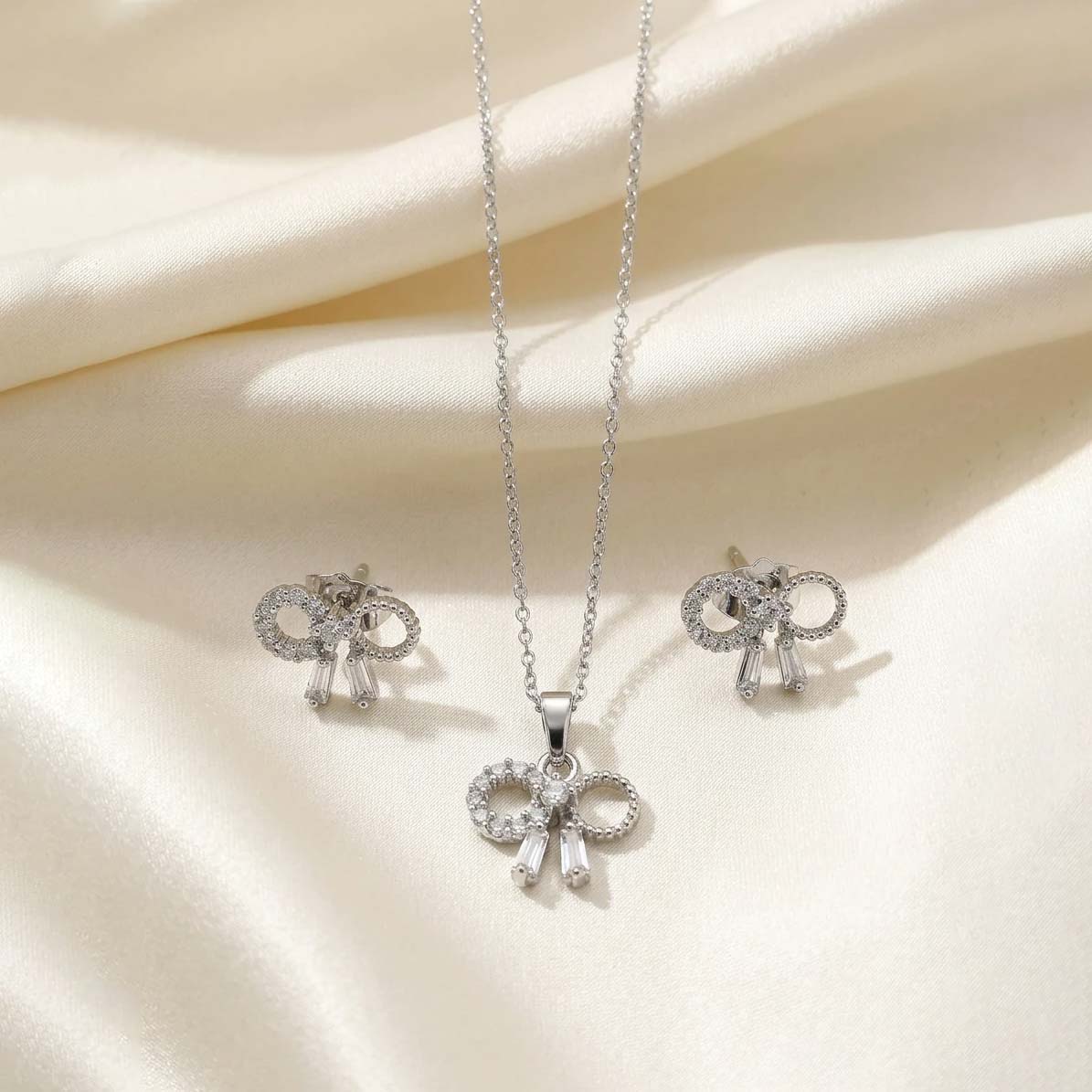 Elegant-Silver-Bow-Jewelry-Set نیمست جواهرات نقرهای رنگ شامل گردنبند ظریف و گوشوارههای میخی با طرح پاپیون نگیندار که روی پارچه حریر روشن قرار گرفته است
