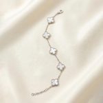 دستبند زنانه نقره‌ای رنگ ظریف دارای پنج آویز طرح گل شبدر چهاربرگ (ون کلیف) با داخل سفید رنگ (صدفی) و دورکاری مهره‌ای ظریف، قرار گرفته روی پارچه ساتن روشن.