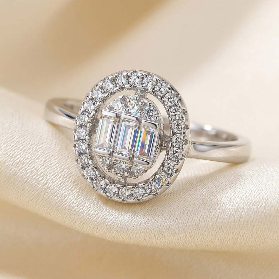 oval-halo-three-baguette-cut-cluster-free-size-engagement-ring انگشتر زنانه فری سایز رنگ نقرهای با طراحی تاج بیضی شکل، دارای سه نگین مستطیلی (تراش باگت) در مرکز که با نگینهای اتمی ریز احاطه شدهاند روی پارچه حریر