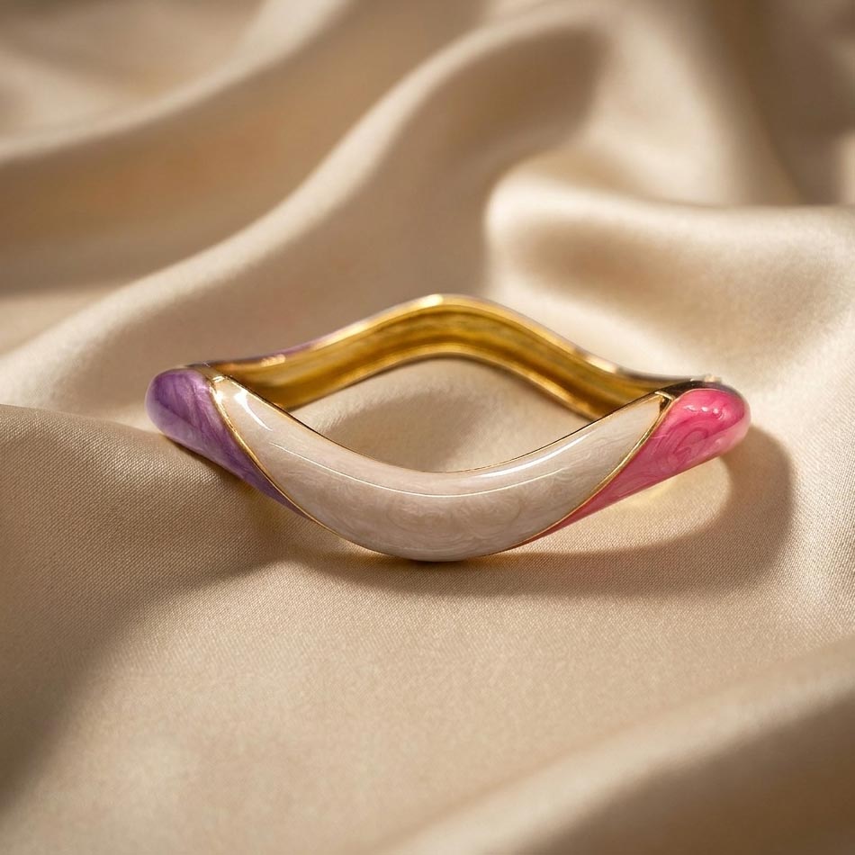Shell-wave-bangle-bracelet دستبند النگویی طرح موج با لبههای طلایی و روکش میناکاری شده به رنگهای بنفش، کرم و صورتی که روی یک پارچه ابریشمی قرار دارد و زیبایی دستبند النگویی طرح موج را نشان میدهد.