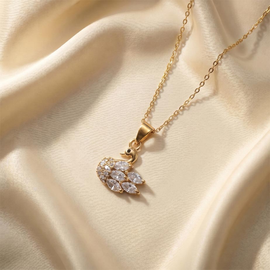 womens-gold-marquise-crystal-swan-necklace گردنبند طلایی رنگ با پلاک طرح قو که پرهای آن با نگینهای اشکی تزئین شده و روی پارچه ساتن کرم رنگ قرار دارد، نمایشی از زیبایی گردنبند طلایی طرح قو.