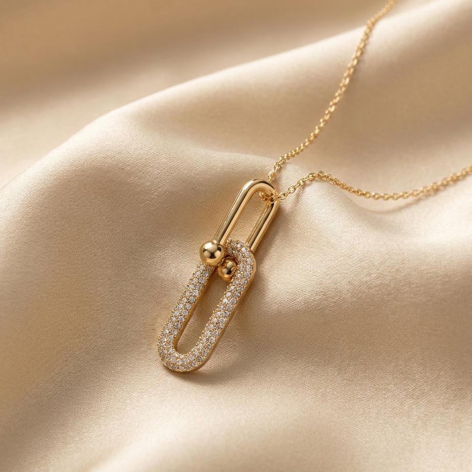 womens-gold-tiffany-style-link-necklace گردنبند طلایی با پلاک متشکل از دو حلقه بیضی متصل به هم که حلقه پایینی با نگینهای ریز تزئین شده و روی پارچه ساتن کرم رنگ قرار دارد، نمایشی از زیبایی گردنبند طلایی طرح تیفانی.