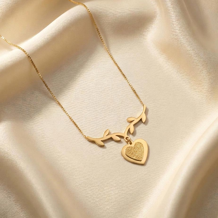 womens-golden-leaf-textured-heart-necklace گردنبند طلایی رنگ با پلاک طرح یک شاخه انحنادار با برگهای کوچک که از مرکز آن یک قلب با سطح داخلی بافتدار آویزان شده و روی پارچه ساتن کرم رنگ قرار دارد، نمایشی از زیبایی گردنبند طلایی طرح شاخه و قلب.
