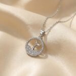 یک گردنبند نقره‌ای با پلاک دایره‌ای که نیمه پایین آن شبیه موج‌های نگین‌دار و وسط آن یک دم وال براق است که روی پارچه ساتن طلایی قرار دارد، نمایش گردنبند طرح دم وال و موج.