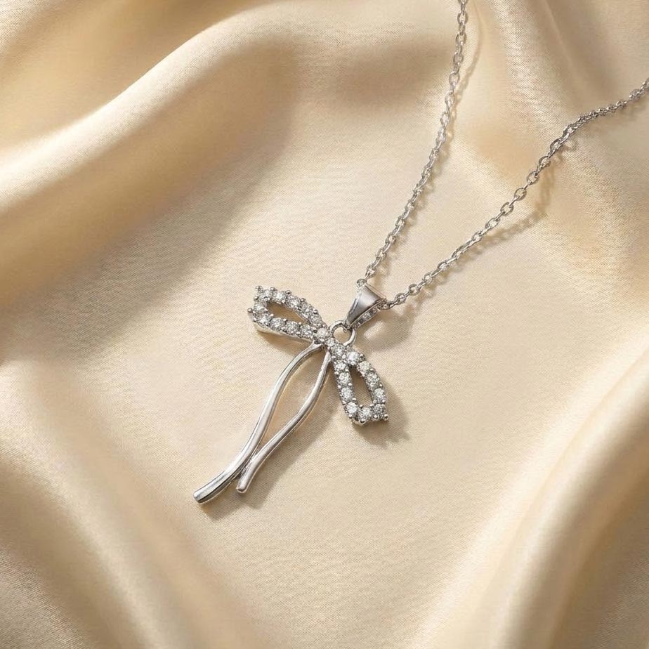 silver-crystal-bow-necklace-long-ribbon تصویر گردنبند طرح پاپیون نقرهای روی پارچه ساتن کرم رنگ که حلقههای پاپیون آن پوشیده از نگین است و دو دنباله صیقلی و بلند از آن آویزان شده است.