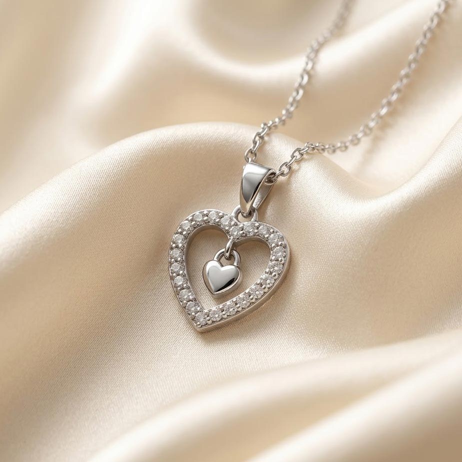 silver-crystal-double-heart-pendant-necklace یک گردنبند طرح دو قلب نقرهای شامل یک قلب بزرگ پوشیده از نگینهای ریز و یک قلب کوچک صیقلی آویزان در مرکز آن که روی پارچه ابریشمی کرم رنگ قرار گرفته است.