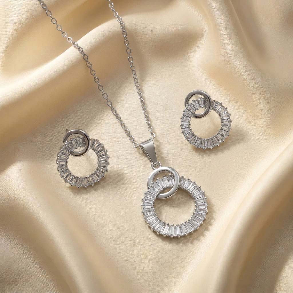 silver-interlocking-circles-baguette-jewelry-set نیم ست نقرهای شامل گردنبند و گوشواره با طرح دو حلقه متصل که یکی از حلقهها پر از نگینهای درخشان باگت است و روی پارچه ساتن کرم قرار دارد.